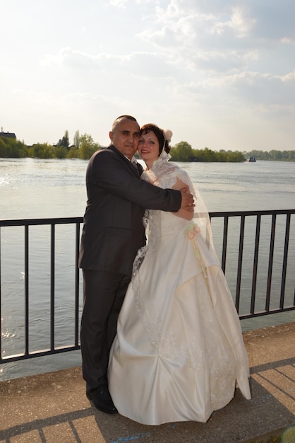 The bride and groom are hugging on the embankment of the river A woman is dressed in a white dress a man in a suit Sunny weather over the river