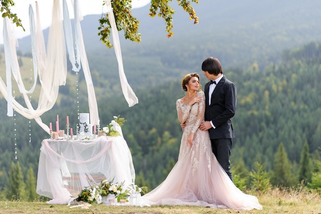 The bride and groom are hugging under an old oak tree. Wedding photo shoot in the mountains. Next to them is prepared decor for the ceremony.