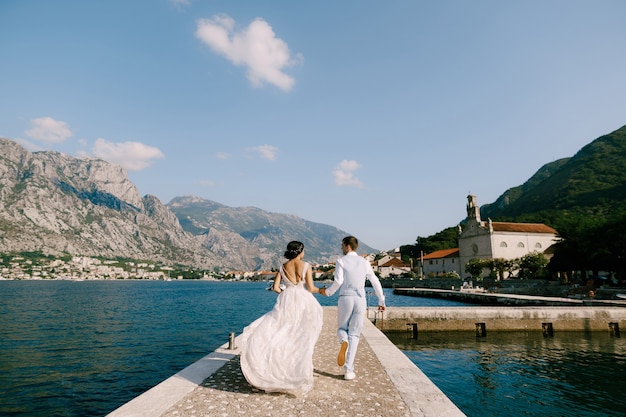 The bride and groom are running along the pier in the bay of kotor holding hands back view