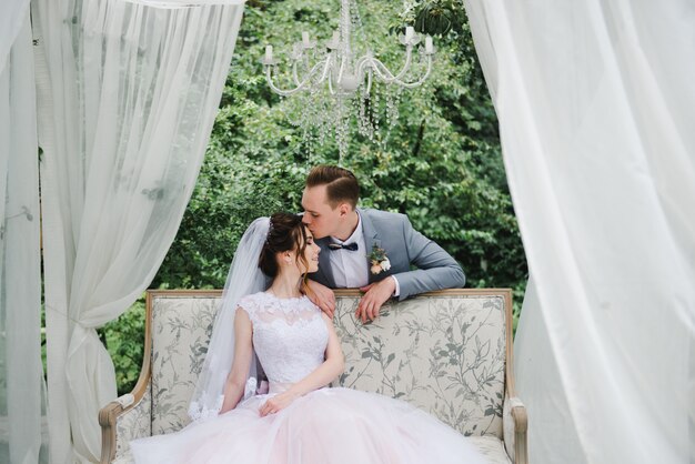 The bride and groom are sitting on a beautiful couch in a gazebo in the garden. 