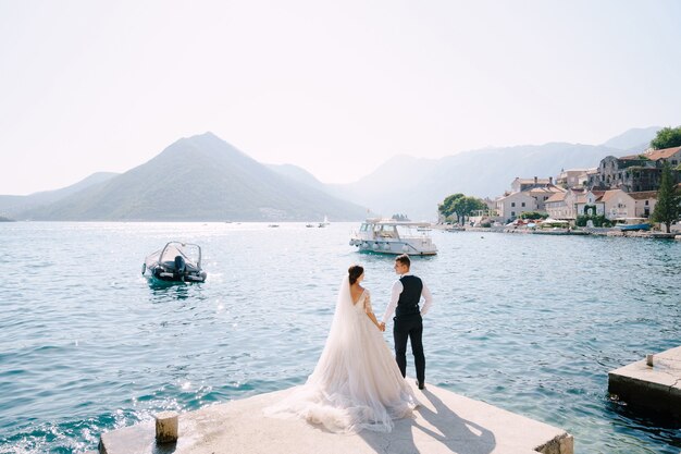 The bride and groom are standing on the pier in the bay of kotor looking at each other against the