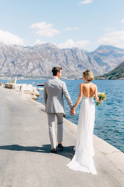 the bride and groom are walking along the pier in the bay of kotor holding hands the bride is