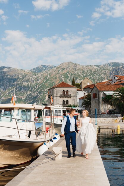 The bride and groom are walking along the pier holding hands in the Bay of Kotor, tourist boats near