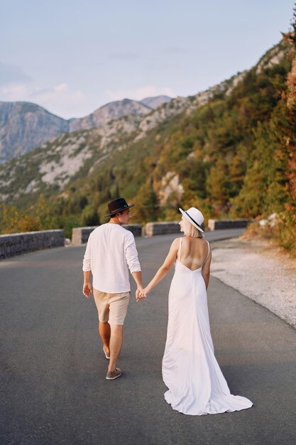 Bride and groom are walking holding hands and looking at each other along the road in the mountains