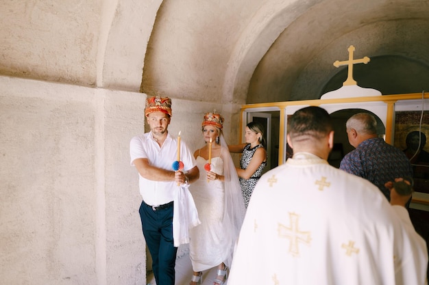 Foto la sposa e lo sposo con le corone e le candele accese nelle mani seguono il prete attraverso la chiesa.