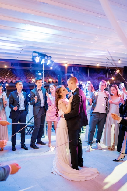 신부 와 신랑 은 댄스 플로어 에서 반 ⁇ 이는 불꽃 을 들고 손님 들 에 둘러싸여  ⁇ 안고 춤 을 추고 있다