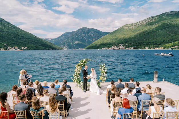 Foto la sposa e lo sposo si tengono per mano all'arco del matrimonio di fronte agli ospiti seduti sul molo