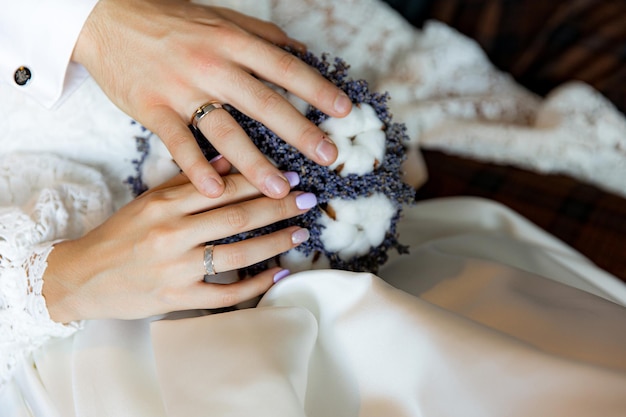 Bride and groom hold hands with rings on a bouquet of lavender with cotton