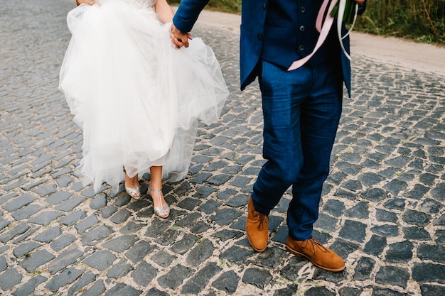 花嫁と花婿は手をつないで、自然の中で道路を歩いて屋外の靴でダウンビュー