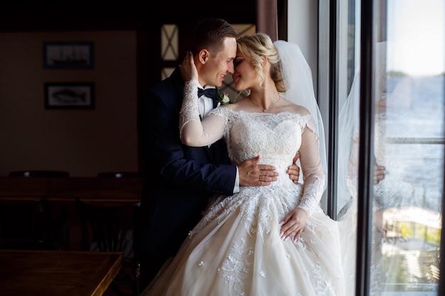 La sposa e lo sposo baci teneramente. foto emotiva di una coppia innamorata il giorno del matrimonio. sposi sorridenti vicino alla grande finestra. foto del matrimonio.