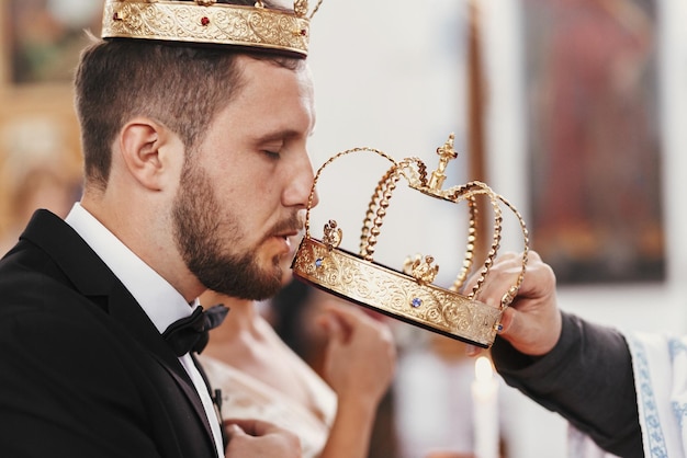 Photo bride and groom kissing golden crowns from priest hand during wedding ceremony spiritual couple wedding matrimony in church emotional romantic moments
