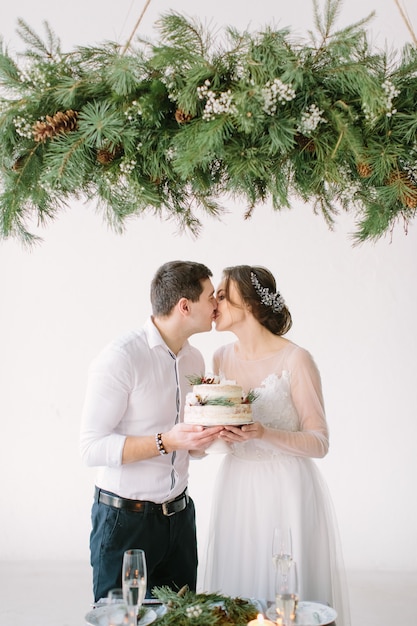 Bride and groom kissing at the table in the banquet hall of the restaurant and holding wedding cake decorated with berries and cotton