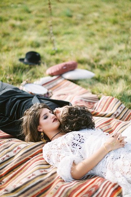 Bride and groom lie on a motley bedspread on a green grass