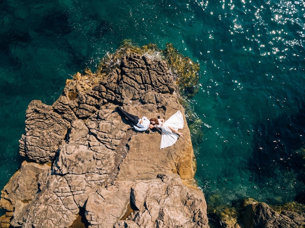 Bride and groom lie on a rocky cliff by the sea aerial view