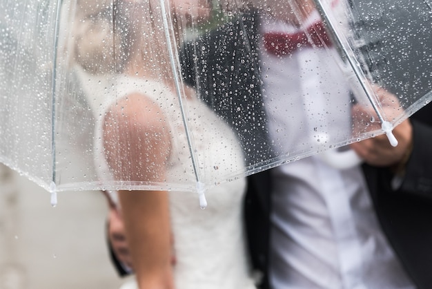 The bride and groom in the rain are covered with a transparent umbrella, rain drops