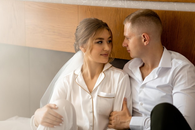 A bride and groom sit on a bed and look at each other.