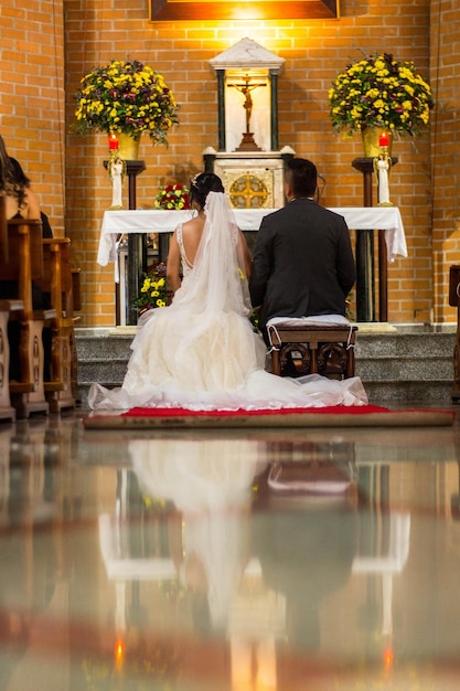 A bride and groom sit in front of a altar with a cross on the altar.