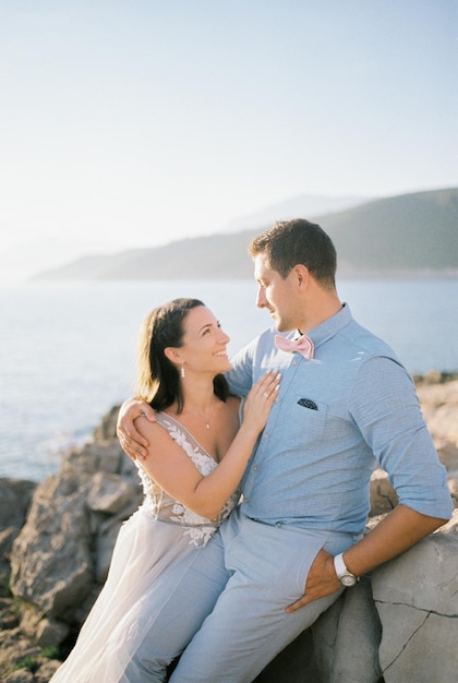 Жених и невеста сидят обнимаясь на скалистом берегу моря