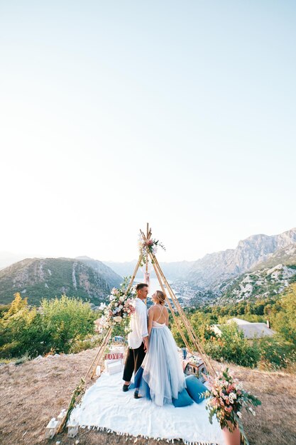 Foto la sposa e lo sposo stanno tenendosi per mano su una coperta sotto l'archwigwam su una montagna sopra la baia di