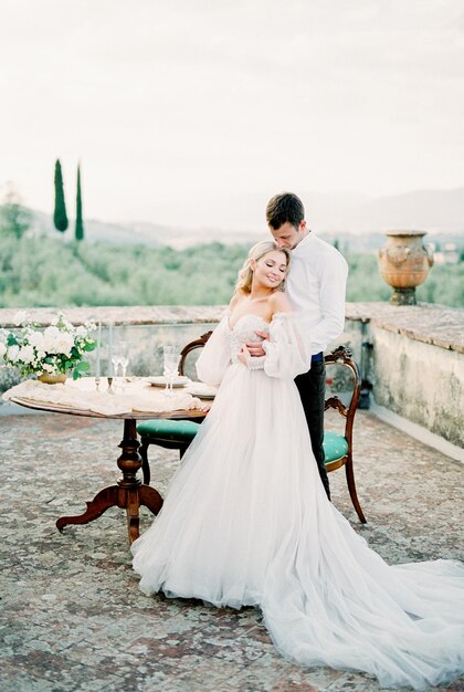 Bride and groom stand near the laid table on the terrace in the garden