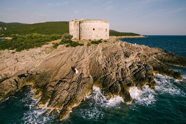 Bride and groom stand on the rocks near the fortress by the sea aerial view