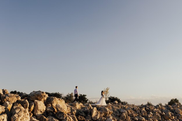 A bride and groom stand on a rocky hill with the sky behind them.