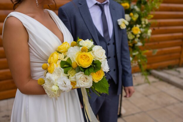Bride and groom standing on green grass and holding a bouquet of white and yellow flowers with green
