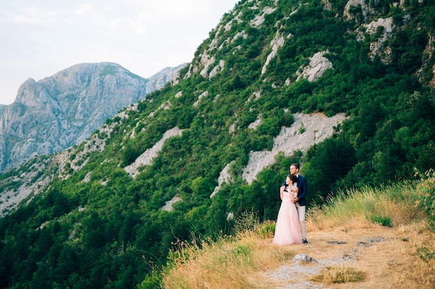 Bride and groom standing on the mountain - the groom gently hugs the bride from behind.