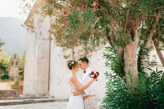 The bride and groom tenderly hugging near the flowering oleander bush near the ancient church in