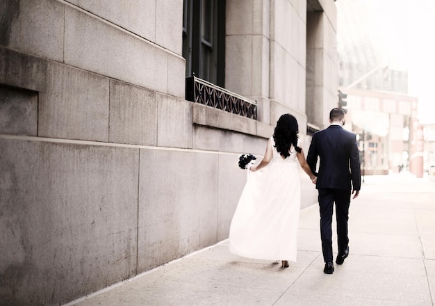 A bride and groom walk down a sidewalk, holding hands, and the groom's dress is wearing a white wedding dress.