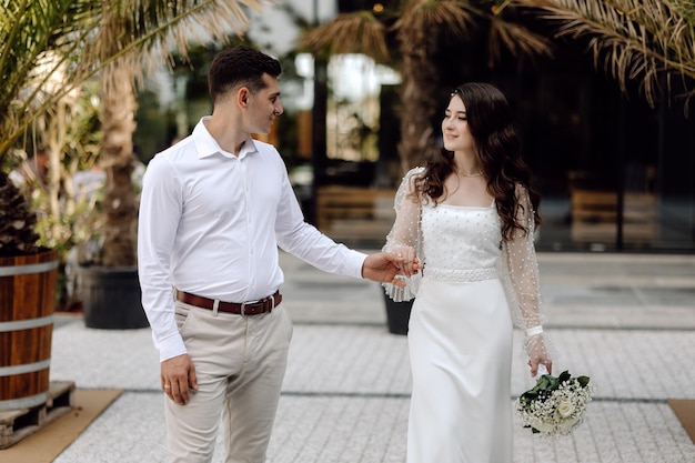 A bride and groom walk hand in hand, the groom wears a white dress with a lace sleeve.