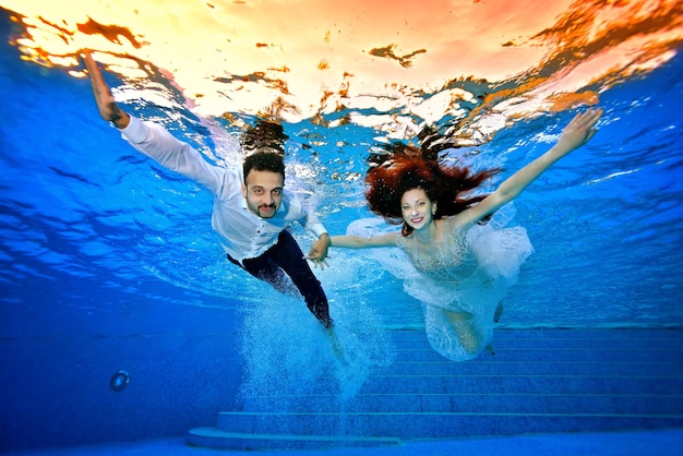 The bride and groom in wedding dresses swim underwater during a bright sunset on the surface