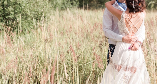 The bride and groom in a white dress are embracing among the tall grass in the meadow