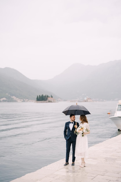 Sposa e sposo con un portaombrelli sul molo in riva al mare sullo sfondo delle montagne