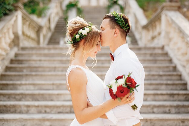 The bride and groom in wreaths stand hugging and kissing on the stairs