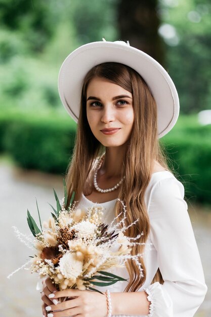 Photo the bride in a hat and a bouquet portrait of a bride in a white dress portrait of the bride young girl in a white wedding dress and hat with a bouquet of flowers