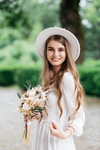 Photo the bride in a hat and a bouquet portrait of a bride in a white dress portrait of the bride young girl in a white wedding dress and hat with a bouquet of flowers