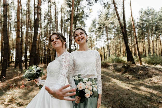 신부와 그녀의 친구는 웨딩 드레스를 입고 손에 꽃다발을 들고 숲에서 즐거운 시간을 보내고 있습니다. 여성의 우정. 결혼식 날. 소녀들은 탐닉하다