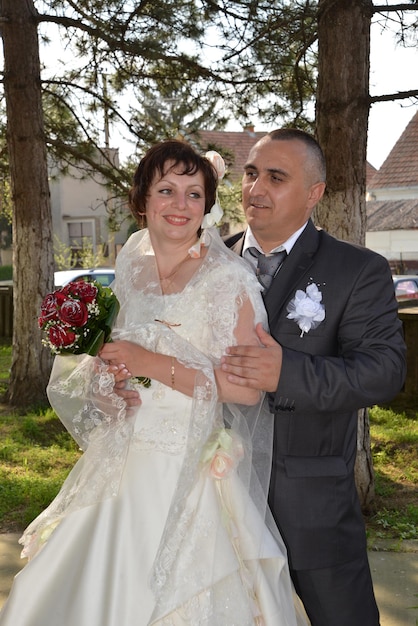 The bride holds a bouquet of red roses in her hands and smiles The groom in a suit looks at the bride and smiles Man holds a womans arm