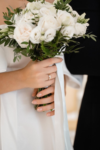 The bride holds in front of her a bouquet of white roses and greenery