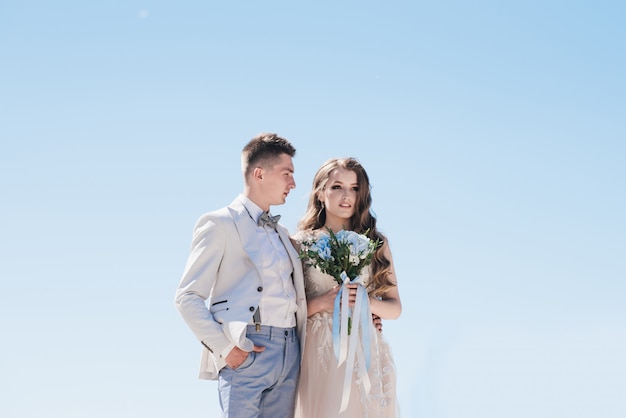 写真 空を背景に軽いスーツで新郎を抱き締める美しいドレスの花嫁。