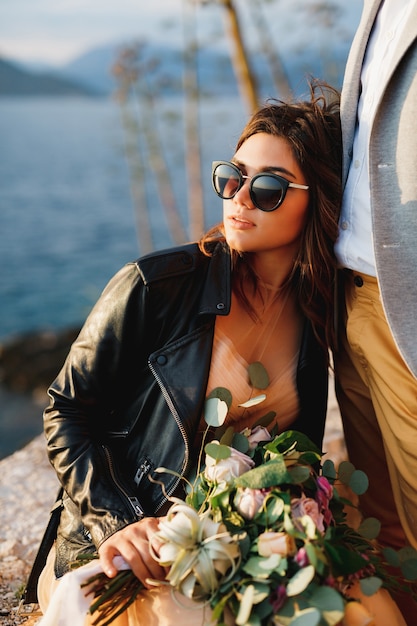 Bride in a leather jacket and sunglasses with a bouquet of flowers is sitting on a stone leaning