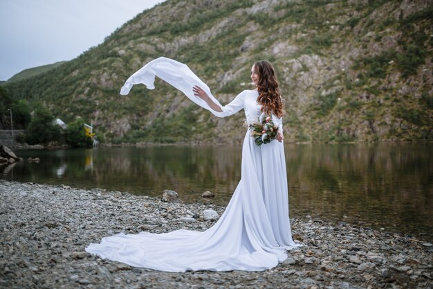a bride in a long wedding dress standing on the shore of a lake, with the sleeves of her dress fluttering
