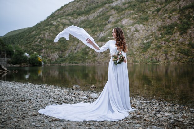 A bride in a long wedding dress standing on the shore of a lake, with the sleeves of her dress fluttering