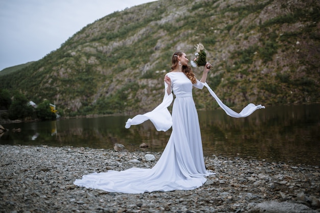 a bride in a long wedding dress standing on the shore of a lake, with the sleeves of her dress fluttering