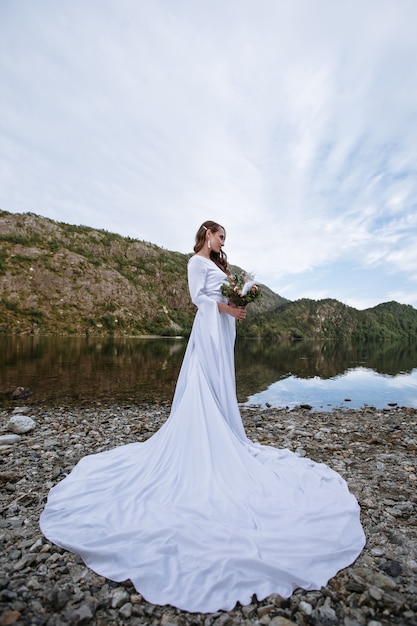 a bride in a long wedding dress standing on the shore of a lake