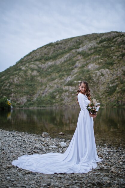 a bride in a long wedding dress standing on the shore of a lake