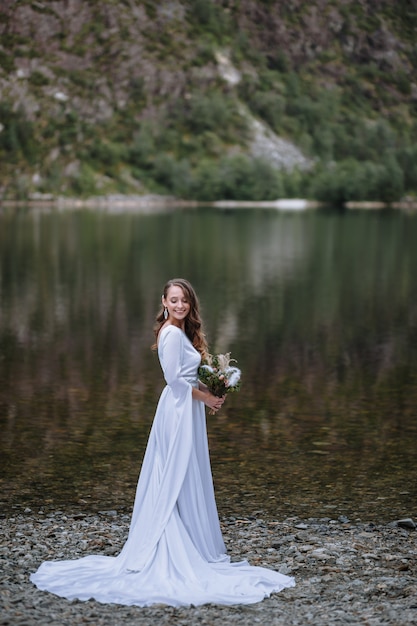 Una sposa in un lungo abito da sposa in piedi sulla riva di un lago