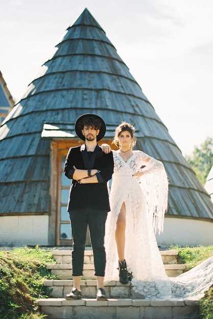 Photo bride put her hand on the shoulder of groom standing on the steps near the conical house