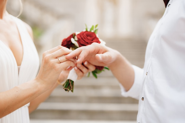 The bride puts a wedding ring on the groom's finger and holds a bouquet of red and white roses